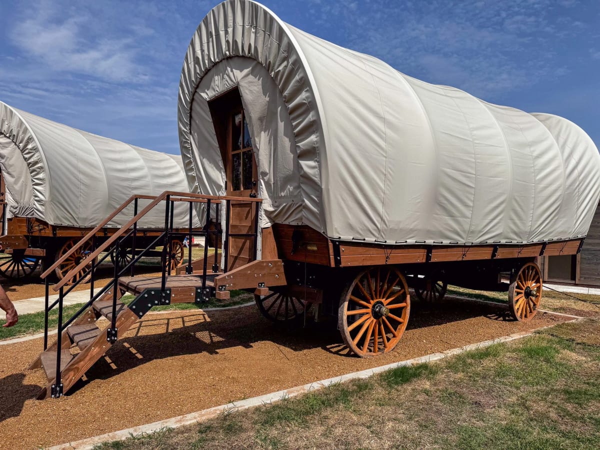 A covered wagon with a white canvas top and wooden base, featuring a small front staircase and large wooden wheels, set outdoors on a dirt and grass surface under a blue sky with clouds.