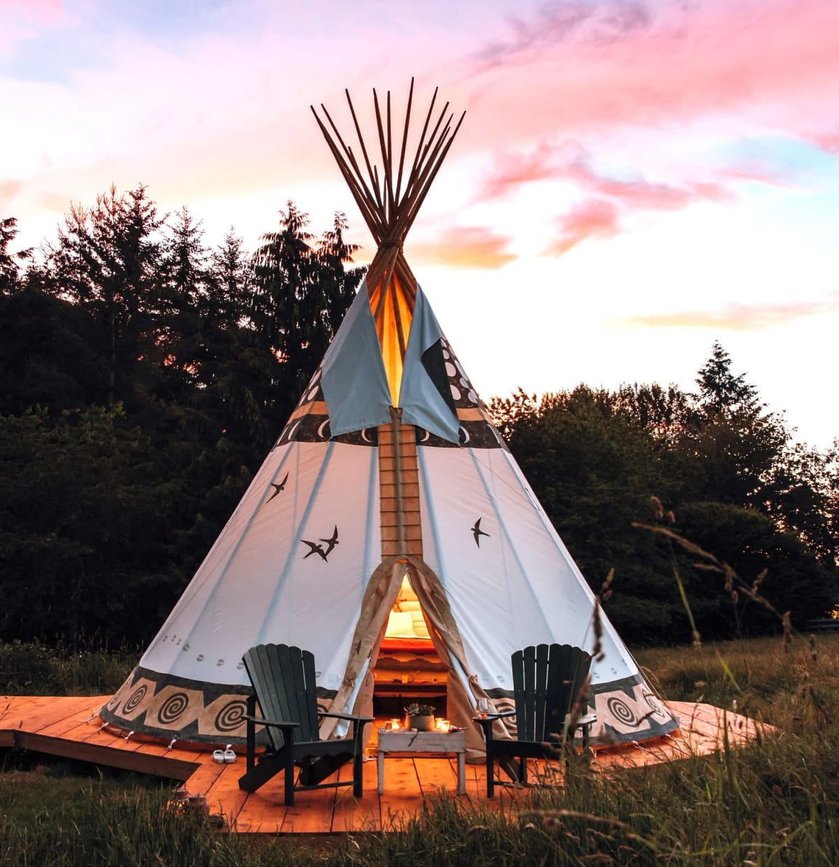 A teepee tent with a decorative design sits on a wooden platform outdoors during sunset, with two chairs, a small table, and a lantern in front.